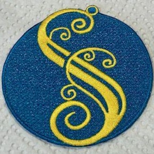 May include: A circular, blue embroidered patch featuring a gold, ornate letter "S". The patch has a textured finish and a small loop at the top for attachment.