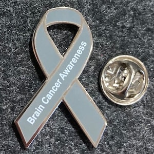 May include: Gray ribbon pin with a silver backing. The ribbon has the words "Brain Cancer Awareness" printed on it.