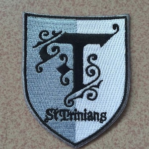 May include: A grey and white embroidered patch with a black letter "T" surrounded by black swirls and the text "St Trinian's".