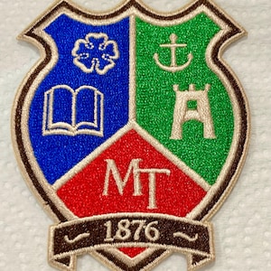 May include: Embroidered shield-shaped patch with blue, green, and red segments. The blue section shows a flower and an open book, the green segment has an anchor and a castle. The red segment has "MT". A banner below reads "1876".