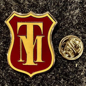 Può includere: Spilla a forma di scudo con sfondo rosso e bordi dorati. La spilla presenta le lettere dorate "TM". Una piccola spilla rotonda color oro è accanto alla spilla a scudo.