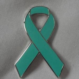 May include: A teal ribbon pin with a silver edge. The ribbon is in a looped shape, often used as a symbol of awareness. The pin is set against a white fabric background.