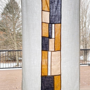 Puede incluir: Una cortina de lino blanco con un panel vertical de tela que presenta un patrón geométrico de cuadrados de color amarillo mostaza, beige y azul oscuro.