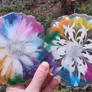 Puede incluir: Dos posavasos circulares de resina con bordes festoneados, que presentan vibrantes patrones florales multicolores en tonos azul, rosa, amarillo, naranja y verde, con un grupo central de purpurina plateada incrustada en el centro de cada pieza.