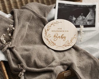 May include: A neutral-toned baby outfit with a wooden announcement disc that reads "Wir Bekommen Ein Baby" in script. The disc is decorated with a floral wreath. An ultrasound image is in the background.