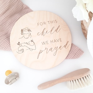 Könnte beinhalten: Ein rundes Holzschild mit einem Laser-geschnittenen Design einer Mutter, die ein Baby hält. Das Schild lautet "Für dieses Kind haben wir gebetet".