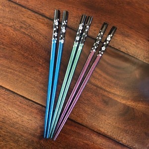 May include: Set of six colorful chopsticks in blue, green, and purple. The chopsticks have black tops with a white floral design. The chopsticks are arranged on a dark wood surface.