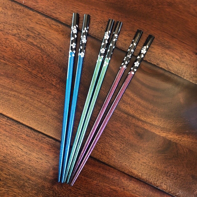May include: Set of six colorful chopsticks in blue, green, and purple. The chopsticks have black tops with a white floral design. The chopsticks are arranged on a dark wood surface.