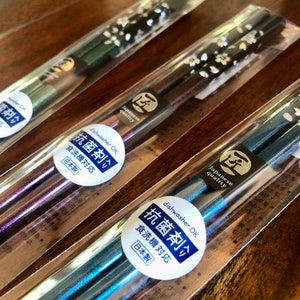 May include: Several pairs of Japanese chopsticks in clear plastic packaging. The chopsticks have a metallic finish in shades of green, purple, and blue. Each pair is decorated with a floral pattern and a label that reads "Japanese quality" and "dishwasher OK".