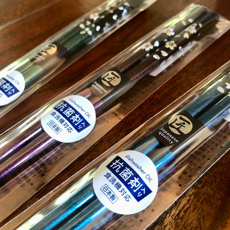 May include: Several pairs of Japanese chopsticks in clear plastic packaging. The chopsticks have a metallic finish in shades of green, purple, and blue. Each pair is decorated with a floral pattern and a label that reads "Japanese quality" and "dishwasher OK".