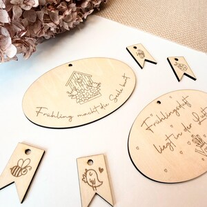 May include: Wooden spring-themed decorations. The image shows several light-colored wooden ovals and pennant shapes with engraved designs. The ovals have text and images of a birdhouse and flowers. The pennants feature a bee, a bird, and a ladybug.