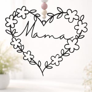 Puede incluir: Decoración de pared en forma de corazón de metal negro con detalles florales y la palabra "Mama" en escritura cursiva. El corazón está adornado con flores y hojas. Una cadena de cuentas blancas, rosas y beige cuelga de la parte superior.