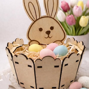 May include: A wooden Easter basket with a bunny cutout on the front. The basket contains colorful eggs and straw. A bouquet of tulips is in the background. The basket is a decorative item for Easter.