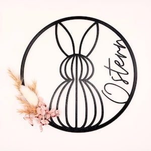 Könnte beinhalten: Schwarze, kreisförmige Osterdekoration mit einer stilisierten Hasen-Silhouette. Der Schriftzug "Ostern" ist kursiv geschrieben. Getrocknete Blumen in Rosa, Beige und Weiß sind auf der linken Seite angebracht.