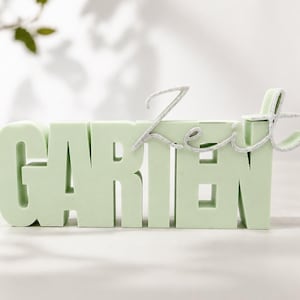 Könnte beinhalten: Ein hellgrünes Schild mit dem Wort "GARTEN" in Blockbuchstaben und einem silbernen, kursiven Overlay mit der Aufschrift "Zeit". Das Schild ist ein dekorativer Gegenstand, möglicherweise für einen Garten. Die Buchstaben sind dick und dreidimensional.