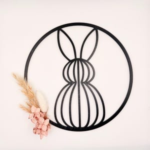 Op de afbeelding: Zwarte metalen wandkunst met een konijnensilhouet in een cirkelvormige lijst. Het konijnontwerp is gestileerd met verticale lijnen. Gedroogde bloemen in beige en roze zijn links onderin geplaatst.