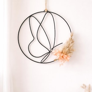 Puede incluir: Adorno de pared de metal negro con forma circular, con un diseño de mariposa formado por líneas curvas. Flores secas en tonos melocotón y beige están unidas al lado derecho. Colgando de un cordel natural. Decoración mural.