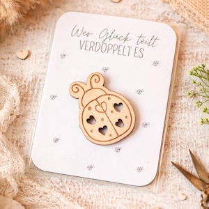 May include: A wooden ladybug-shaped embellishment with heart cutouts is affixed to a white card. The card has the German text "Wer Glück teilt verdoppelt es" and small ladybug illustrations. Small wooden hearts and scissors are also visible.