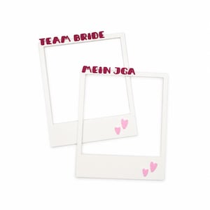 Könnte beinhalten: Zwei weiße, rechteckige Fotorahmen im Design von Sofortbildern. Ein Rahmen trägt oben den dunkelrosa Schriftzug "TEAM BRIDE", der andere "MEIN JGA" in derselben Farbe, verziert mit kleinen hellrosa Herzen auf der unteren weißen Fläche.