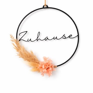 Op de afbeelding: Een zwarte metalen hoepel met het woord "Zuhause" in cursief schrift. De hoepel is versierd met gedroogde beige grassen en perzikroze bloemen. Een natuurlijk touw wordt gebruikt om het bloemstuk te bevestigen.