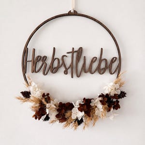 Può includere: Una decorazione da parete circolare in legno scuro con la scritta "Herbstliebe" in corsivo. La metà inferiore del cerchio è decorata con un insieme di petali di ortensia bianchi essiccati, foglie marrone scuro e spighe di grano.