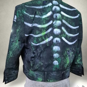 Könnte beinhalten: Eine schwarze Jacke mit einem handgemalten weißen Brustkorb-Motiv auf der Rückseite. Der Stoff hat ein abgenutztes, verwittertes Aussehen mit gesprenkelten grünen Farbflecken. Die Jacke verfügt über einen Kragen, lange Ärmel und einen Saumgürtel.
