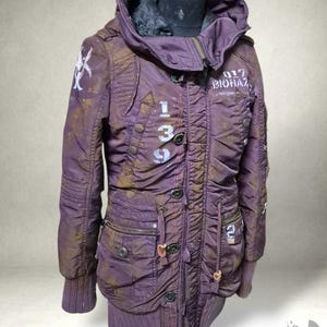 May include: A purple jacket with a hood, multiple pockets, and a drawstring waist. The jacket features white stenciled numbers and text, along with brown accents. The jacket is displayed on a mannequin.