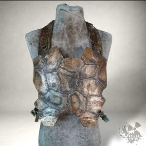 Op de afbeelding: Een verweerd, handgemaakt borstpantser gemaakt van onregelmatig gevormde, metaalachtige platen. Het pantser is voornamelijk bruin en grijs, met zichtbare naden en een textuur. Het heeft schouderbanden en wordt op een mannequin getoond.