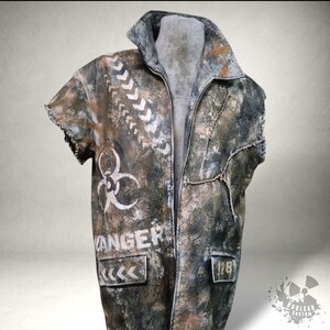 Op de afbeelding: Een distressed jas in post-apocalyptische stijl met een verweerde uitstraling. Het heeft witte chevron- en biohazard-symbolen en het woord "DANGER". Het kleurenpalet omvat bruin, grijs en wit. Met ritssluiting en zakken.