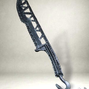 Könnte beinhalten: Ein handgefertigtes Messer aus schwarzem Metall mit einzigartigem Design. Die Klinge hat geometrische Aussparungen und eine gezahnte Kante. Der Griff hat ein hakenförmiges Ende. Das Messer ist ein dekorativer Gegenstand.