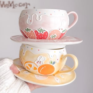 Könnte beinhalten: Zwei Keramik-Teetassen mit passenden Untertassen. Die obere Tasse ist rosa mit Erdbeermotiven, und die untere Tasse ist gelb mit einem Orangenscheiben- und Hasenmuster. Beide Tassen haben einen weißen, tropfenden Glanzeffekt.