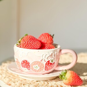 Op de afbeelding: Een roze keramische mok met een aardbeidesontwerp en een schotel. De mok is gevuld met verse aardbeien.