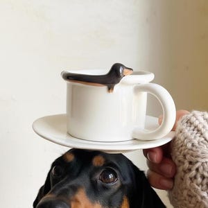 May include: A white ceramic coffee mug with a dachshund dog design on the rim, sitting on a matching saucer. The dog is black and brown. The mug has a large, curved handle.