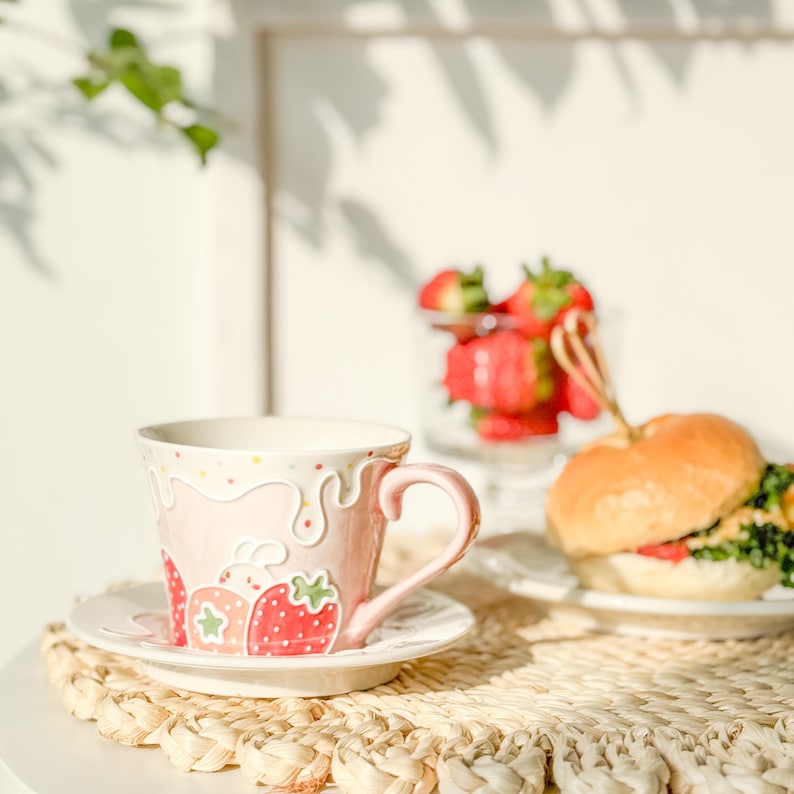 Op de afbeelding: Een roze en witte keramische theekop met een aardbeienontwerp. De theekop staat op een wit schoteltje en een geweven placemat. Er is een bord met een hamburger op de achtergrond.