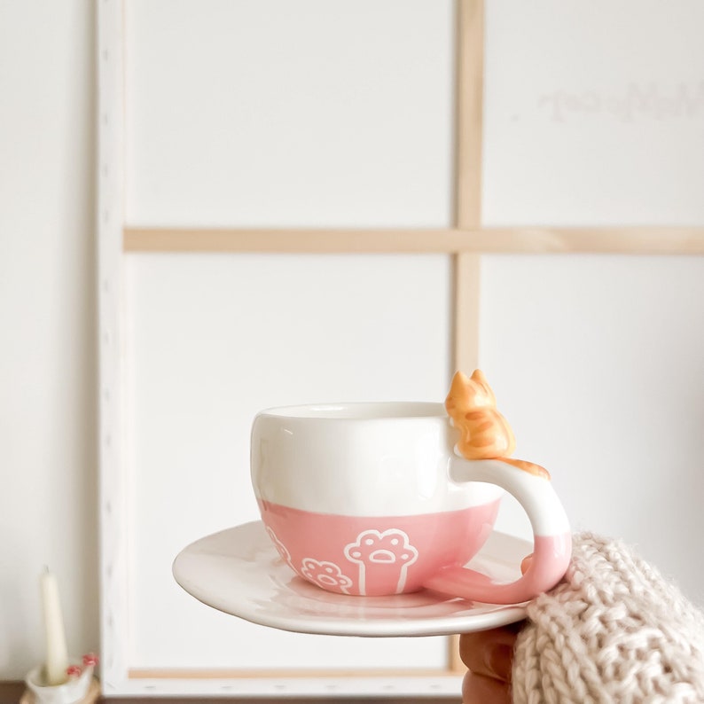 May include: A pink and white ceramic teacup with a cat-shaped handle. The cat is orange and white and is sitting on the rim of the cup. The cup is on a white saucer.