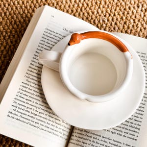 May include: White ceramic teacup and saucer set with a dachshund-shaped handle, resting on an open book. The dachshund handle is orange and white. The book pages are visible, with text in black.