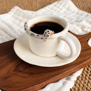 May include: A white ceramic coffee cup and saucer set filled with dark coffee. The cup features a decorative dachshund dog handle with black spots and an orange nose. The set is placed on a wooden tray.