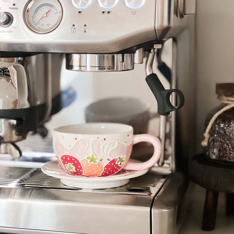 Op de afbeelding: Een espressomachine van roestvrij staal met een kopje koffie op een roze schotel. Het kopje is versierd met aardbeien en een roze konijnenontwerp.