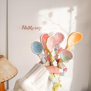 May include: A set of six pastel-coloured ceramic spoons with floral designs and a frog charm. The spoons are decorated with flowers and leaves in shades of pink, blue, yellow, and green.