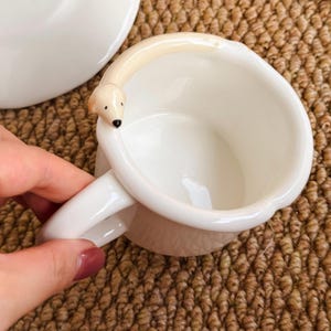 May include: White ceramic mug with a polar bear design on the rim. The mug has a curved handle and a matching saucer is visible in the background. The polar bear has a black nose and small, dark eyes.