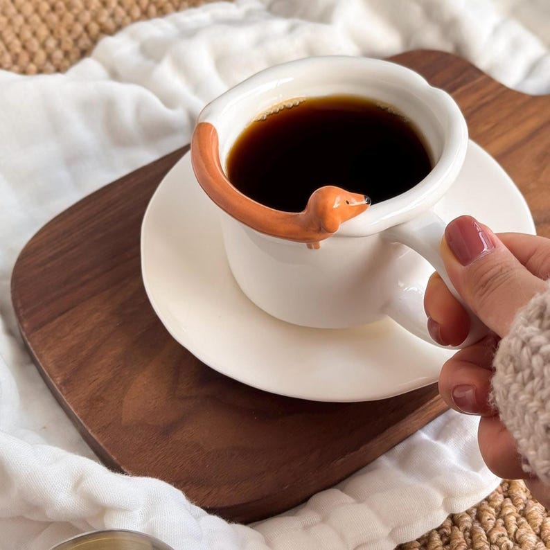 May include: A white ceramic coffee cup and saucer set with a dachshund dog design. The dog's body forms the handle, and its head extends over the cup's rim. The cup is filled with dark coffee, and the set sits on a wooden tray.
