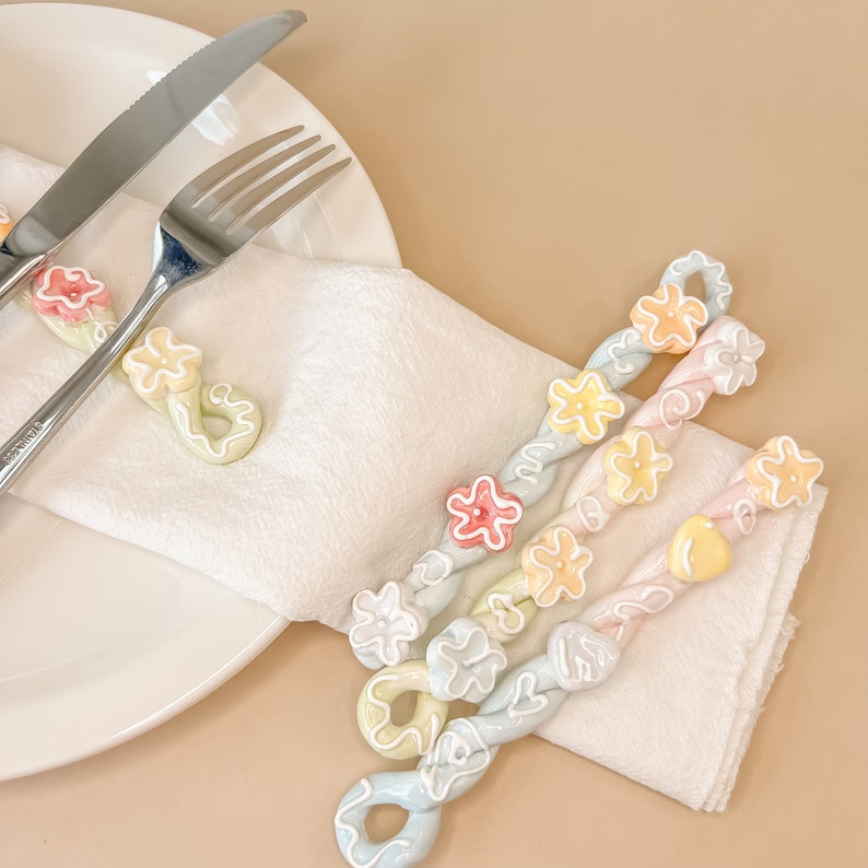 May include: Three pastel-colored napkin rings with flower accents. The rings are made of twisted ceramic and feature a variety of pastel colors, including pink, blue, yellow, and green. The rings are arranged on a white napkin.