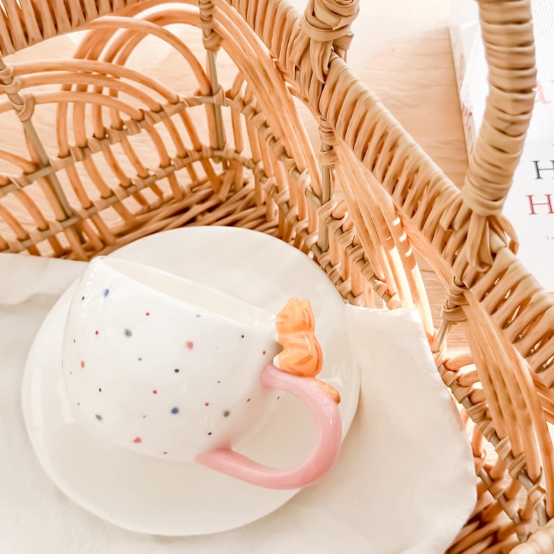 May include: A white ceramic teacup with a pink handle and colorful polka dots. A small squirrel figurine is attached to the side of the cup. The cup and saucer are sitting inside a wicker basket.