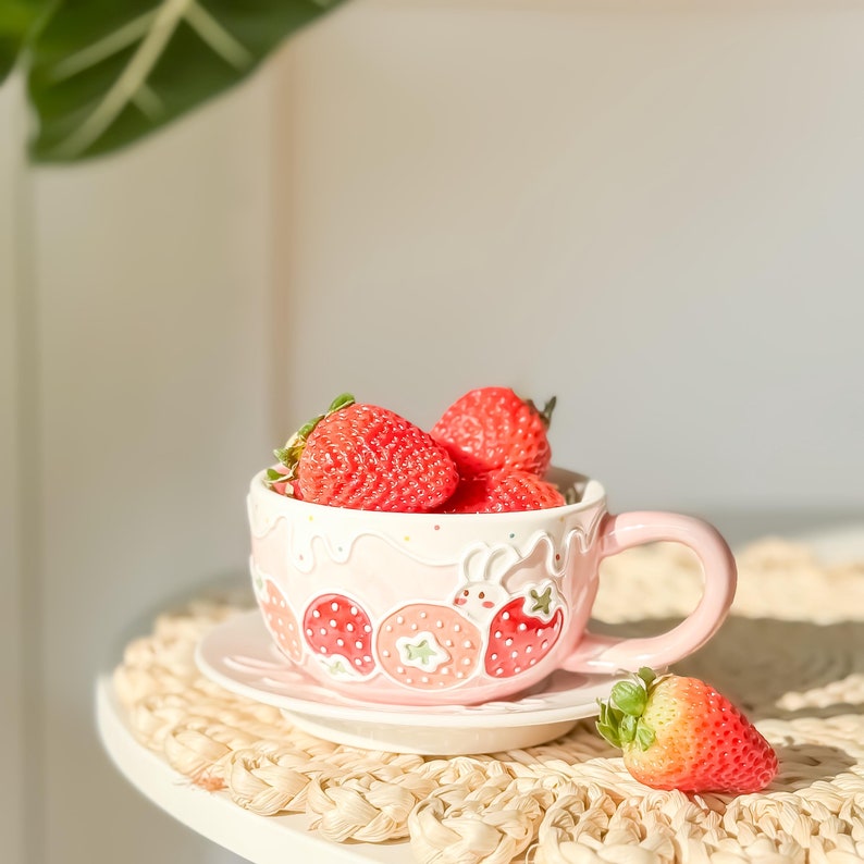 Op de afbeelding: Een roze keramische mok met een aardbeidesontwerp en een schotel. De mok is gevuld met verse aardbeien.