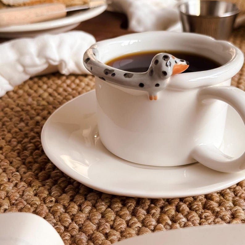May include: White ceramic coffee cup and saucer set. The cup is filled with dark coffee and features a decorative dachshund dog design on the rim. The dog is gray with black spots and an orange nose.