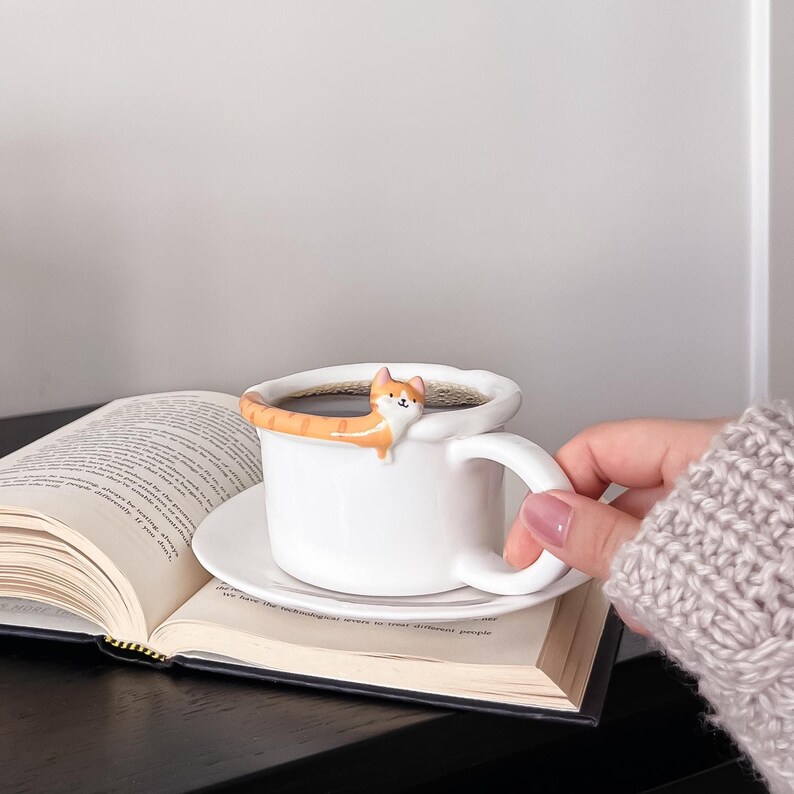 May include: A white ceramic coffee mug with a small orange cat figurine perched on the rim. The cat's tail is curled around the mug. The mug is sitting on a white saucer on top of an open book.
