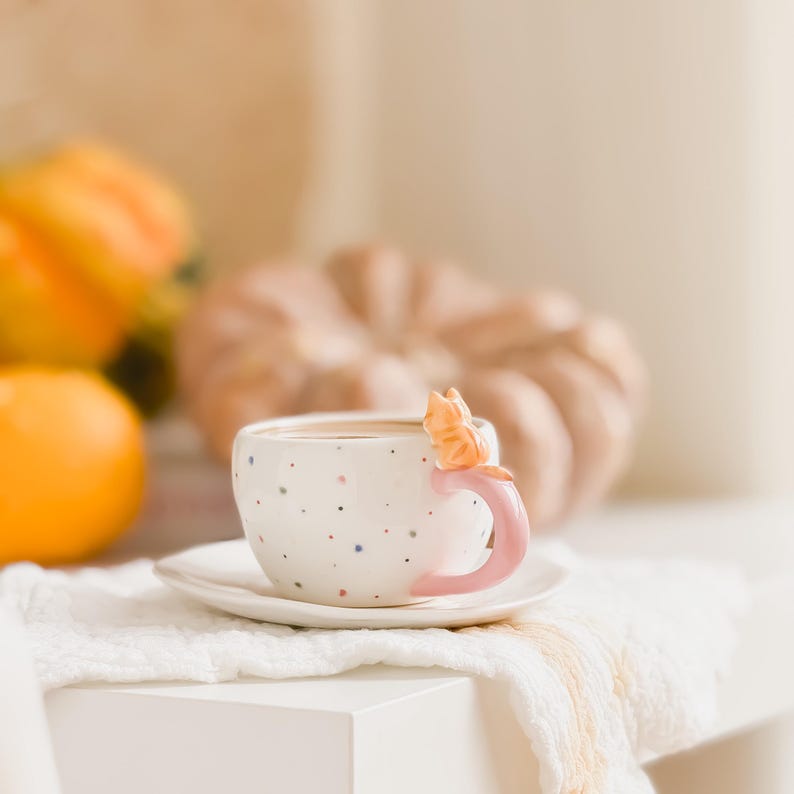 May include: A white ceramic teacup with a pink handle and a small orange cat figurine on the side. The cup is filled with a light brown liquid and is sitting on a white saucer. The cup is on a white table with a white cloth napkin.