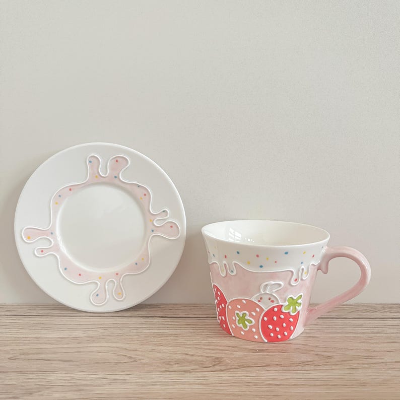 Op de afbeelding: Een wit theeservies met een roze en witte glazuur. De theekop heeft een handvat en een roze en witte glazuur met een aardbeienontwerp. Het schoteltje heeft een roze en witte glazuur met een wit splatterontwerp.