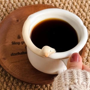 May include: A white ceramic coffee mug filled with dark liquid, featuring a dachshund-shaped handle. The mug sits on a wooden coaster with text. The mug is held by a person wearing a cream-colored sweater.
