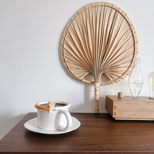 May include: A white ceramic coffee mug with a brown cat figurine perched on the rim. The mug is filled with coffee and is sitting on a white saucer. The mug is on a dark brown wooden table. A large, beige palm leaf fan is on the wall behind the mug.